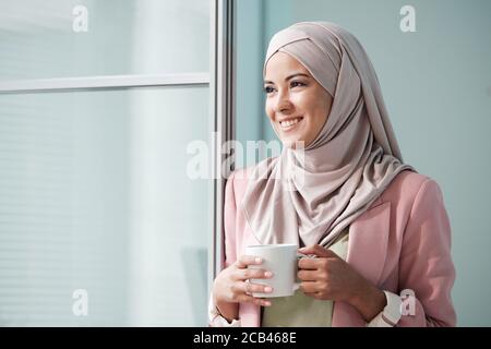 Allegro attraente giovane donna musulmana in rosa hijab in piedi a. porta e tenendo la tazza mentre si guarda via Foto Stock
