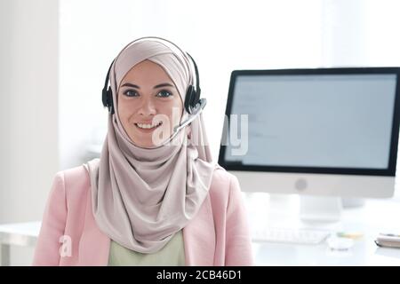 Ritratto di bella donna musulmana positiva in risposta hijab domande sull'utilizzo del dispositivo vivavoce nel call center Foto Stock
