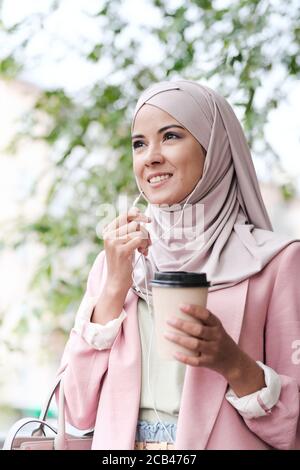 Sotto la vista di sorridente giovane e fiduciosa donna d'affari araba in hijab bere caffè e utilizzare gli auricolari mentre si risponde al telefono in movimento Foto Stock