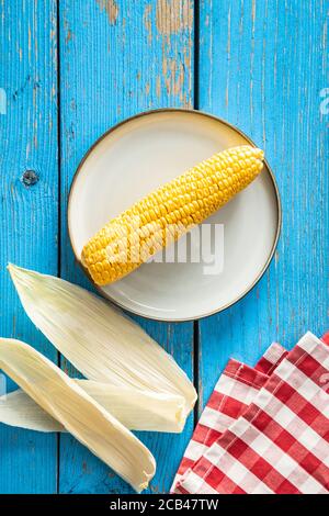 Pannocchie di mais non cotte su piastra su tavola di legno blu. Vista dall'alto. Foto Stock