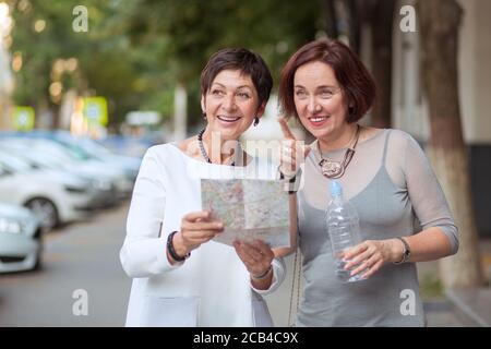 Elegante donna di mezza età con bottiglia d'acqua e mappa in piedi sulla strada in cerca di direzione mentre si viaggia insieme Foto Stock