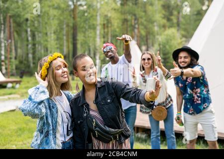 Donna nera positiva con lunghi orecchini che fotografano con un amico wreath sullo smartphone mentre i loro amici fanno i gesti cool dentro sfondo Foto Stock