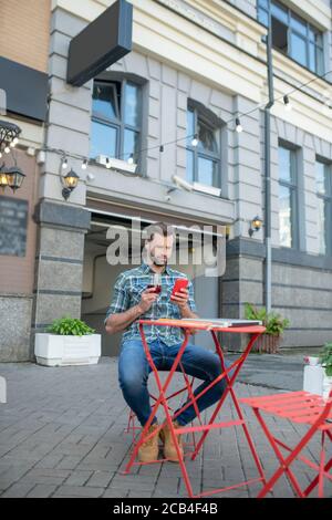 Bearded bel maschio che beve vino rosso, controllando il suo telefono in un caffè all'aperto Foto Stock