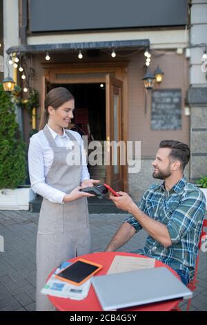 Pagando il conto maschile con smartphone al bar all'aperto, cameriera che tiene il terminale di pagamento Foto Stock