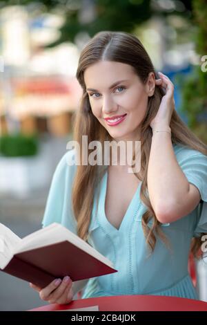 Affascinante femmina dai capelli castani, libro di lettura, stropicciando i capelli, sorridendo Foto Stock