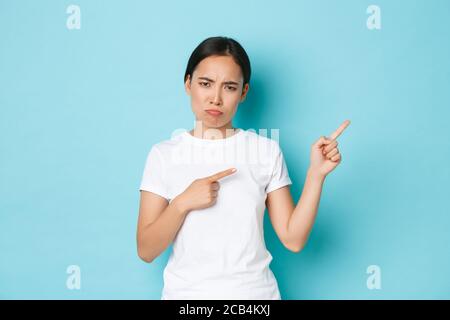 Deluso triste sulking asian ragazza in bianco t-shirt accigliata e aspetto fastidioso, puntando le dita in alto a destra, lamentando qualcosa di ingiusto Foto Stock