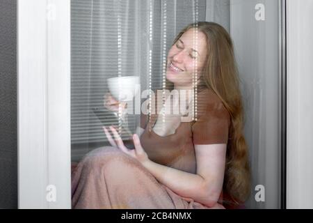 una ragazza si siede su un davanzale vicino alla finestra, avvolta in una coperta, con una tazza bianca per il tè e tiene una tavoletta in mano, con gli occhi chiusi gioiosamente en Foto Stock