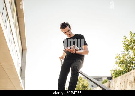 Uomo d'affari seduto sulla ringhiera e che utilizza un tablet digitale fuori dall'ufficio contro il cielo Foto Stock