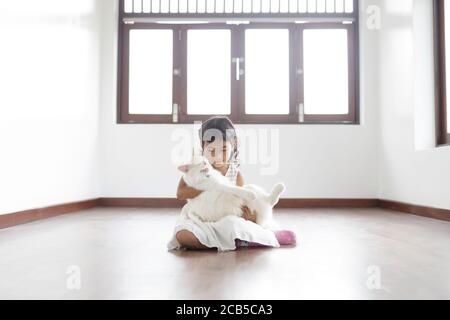 Una ragazza asiatica carina sta tenendo il suo gatto bianco seduto sul pavimento all'interno di una casa dipinta di bianco. Foto Stock