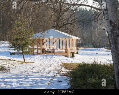 grande gazebo di alcova in legno con panca nella neve del giardino d'inverno betulla e abete rosso al sole del pomeriggio Foto Stock