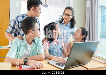 Gruppo di studenti adolescenti asiatici e il loro insegnante di informatica si è riunito alla scrivania con il laptop per discutere il programma del computer Foto Stock