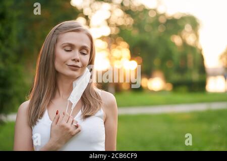 Vista frontale di una giovane donna sbalorditiva con gli occhi chiusi togliendo la maschera protettiva bianca. Bella bionda femminile in posa in verde parco, bellissimo tramonto estivo sullo sfondo. Concetto pandemico. Foto Stock