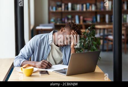 Un uomo nero esausto che studia online su un computer portatile in un ambiente accogliente caffetteria Foto Stock