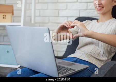 Datazione online. Vista ritagliata della giovane donna che mostra il cuore con le mani al suo ragazzo mentre parla online sul portatile Foto Stock