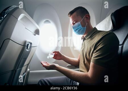 Uomo che indossa la maschera facciale e usa l'igienizzatore per le mani all'interno dell'aereo durante il volo. Temi nuovo normale, coronavirus e protezione personale. Foto Stock