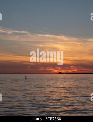 La silhouette di un uomo in piedi su una tavola da surf sul mare come il sole tramonta sul nucleare Centrale elettrica Barsebäck vista in lontananza Foto Stock