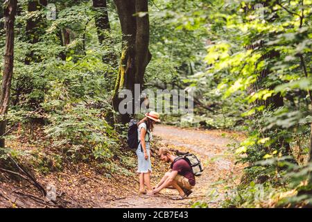 Il giovane caucasico con la barba si prende cura della donna, legando i lacci sulle sue scarpe mentre cammina nella foresta lungo il sentiero. Coppia di escursionisti con Foto Stock