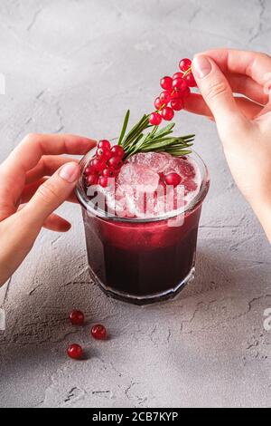 Le mani della donna contengono bacche di ribes per la preparazione di cocktail di frutta fresca ghiacciata in vetro con foglia di rosmarino, fondo di pietra in cemento Foto Stock