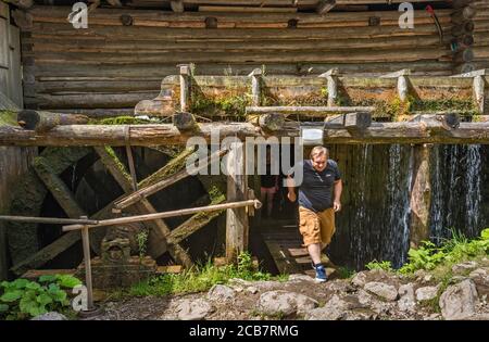 Turista che esce sotto il fumo, acqua che perde, ruota dell'acqua di colpo in eccesso presso lo storico mulino ad acqua di Oblazy al fiume Kvacianka, Valle di Kvacany (Kvačianska dolina), zona di Liptov, Regione di Zilina, Slovacchia Foto Stock