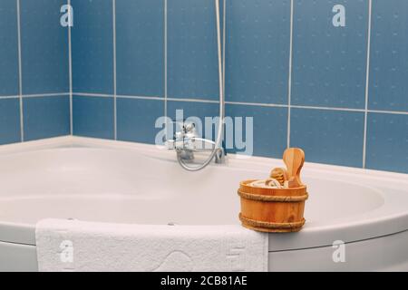 Mestolo di legno con spazzola, panno e altri accessori doccia sul bordo di una vasca ad angolo vicino a un asciugamano terry su uno sfondo di piastrelle blu Foto Stock