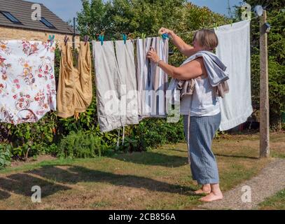 Una donna più anziana che disinfetta e raccoglie una linea di lavaggio a secco su una linea esterna in un giardino / cortile, sotto il sole forte. Foto Stock