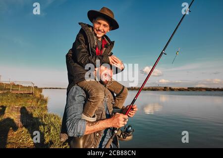 Figlio e padre si divertono mentre pesca. Padre tiene il figlio sulle spalle. Ragazzo tenere testa. Sorridono. Sfondo lago. Foto Stock