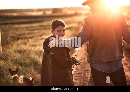 Padre e figlio hanno preso canne da pesca e attrezzatura da pesca. Vanno sulla strada di campagna vicino al lago per la pesca. Tramonto. Foto Stock