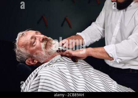 vecchio uomo bearded sdraiato sulla sedia con gli occhi chiusi che ottengono il taglio dei capelli. primo piano della foto Foto Stock