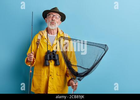 divertente vecchio uomo con binoculare, asta e rete in giallo raincaot e grren cappello che soffia bacio, fischio primo piano foto. isolato sfondo blu, studio sh Foto Stock