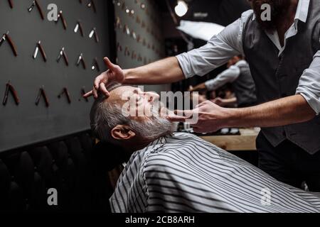 Senior rispettabile uomo caucasico visitando un parrucchiere in un barbiere per la rasatura e la creazione di un nuovo design per il suo grigio dai capelli barba Foto Stock