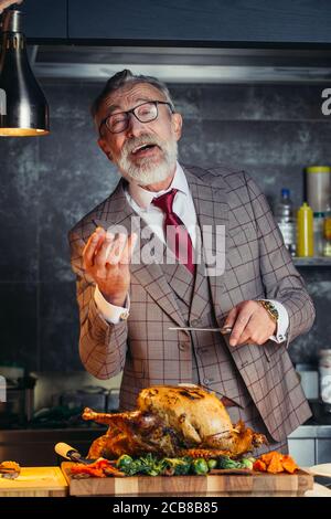 Il vecchio uomo vestito di ricco vestito su misura carving deliziosi carbone succulento pollo arrosto con coltello su un tagliere di legno, vacanze in famiglia e cel Foto Stock