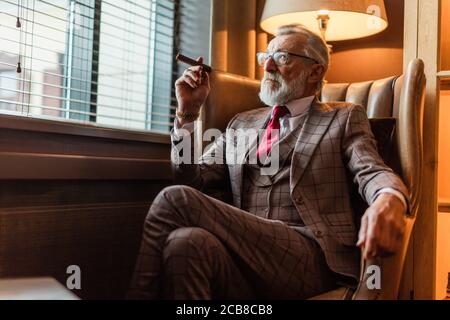 Grave di vecchia età imprenditore indossando elegante personalizzato suit, cravatta rossa e costoso orologio da polso seduto sul vecchio divano in ufficio, guardando Foto Stock