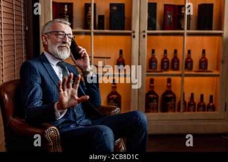 Vecchio uomo d'affari riuscito in abbigliamento costoso formale che parla sullo smartphone con il suo cliente, gesturing NO al cameriere portando il susino seguente, sedendo io Foto Stock