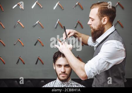 Processo di lavoro in barbiere moderna. Parrucchiere maschile che serve il cliente, facendo taglio di capelli con forbici e pettine di metallo, vista laterale. Foto Stock