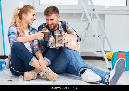 Giovane famiglia di decoratori di casa che si raffredda in camera vuota preparata per la pittura, seduto tra gli attrezzi, che sono sempre necessari durante il tempo di riparazione instru Foto Stock