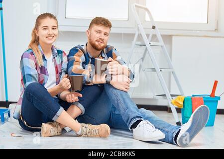 Giovane famiglia di decoratori di casa che si raffredda in camera vuota preparata per la pittura, seduto tra gli attrezzi, che sono sempre necessari durante il tempo di riparazione instru Foto Stock