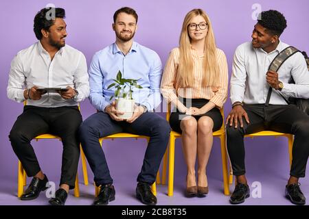 giovani di diverse nazionalità seduti di fila atmosfera amichevole isolato su viola in attesa di colloquio di lavoro Foto Stock