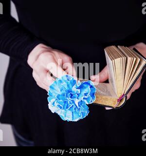 Mani femminili che tengono il vecchio libro e fiore blu di garofano su uno sfondo nero. Spazio di copia Foto Stock