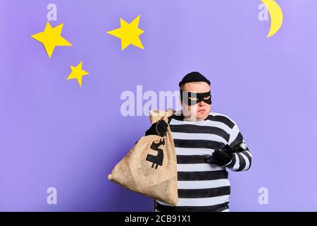 ladro grasso serio che porta una borsa grande, vestita con abiti a righe, maschera e guanti che tengono una torcia di notte. primo piano foto. sfondo blu isolato Foto Stock