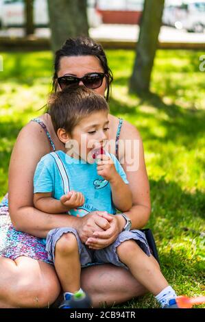 POZNAN, POLONIA - 26 luglio 2020: Donna che indossa occhiali da sole seduto con un bambino su un campo di erba verde in una calda giornata estiva. Ragazzo sta mangiando ghiaccio di fragola Foto Stock