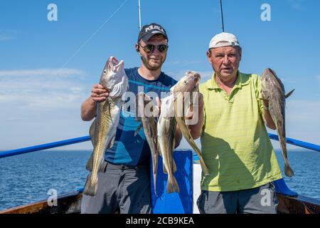 Malmo, Svezia - 6 agosto 2020: Due uomini in un viaggio di pesca mostrano la loro cattura fresca di merluzzo. Oceano blu e cielo sullo sfondo Foto Stock