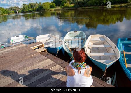 Una donna si siede su un molo di legno e ammira il fiume e le barche. Foto Stock