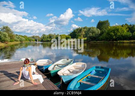 Una donna si siede su un molo di legno e ammira il fiume e le barche. Foto Stock