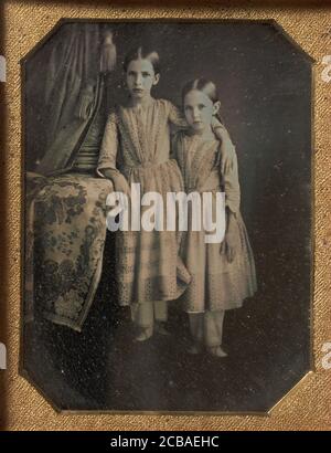 Due ragazze giovani vestiti in modo identico che si levano in piedi vicino ad un tavolo, 1840. Foto Stock