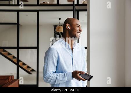 Foto di un gioioso uomo afro-americano che ride mentre usa il wireless auricolare e cellulare in soggiorno Foto Stock