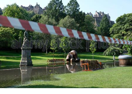 Edimburgo, Scozia, Regno Unito. 12 agosto 2020. Il tempo soleggiato sostituisce le tempeste notturne che lasciano i West Princes Street Gardens allagati e cordonati al pubblico. Scultura in bronzo elefante intrappolata nelle acque alluvionali. Vista sul Castello di Edimburgo. Credit: Craig Brown/Alamy Live News Foto Stock