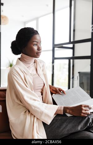 Foto di giovane donna africana piuttosto concentrata seduta sul divano in interni a casa durante la lettura di riviste o libri Foto Stock