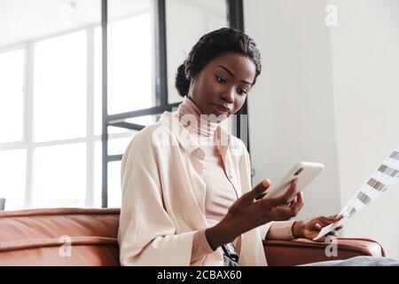 Attraente giovane donna africana seduta su un divano a casa, interni moderni, la scelta di nuovi colori per la ristrutturazione della casa, tenere telefono cellulare Foto Stock