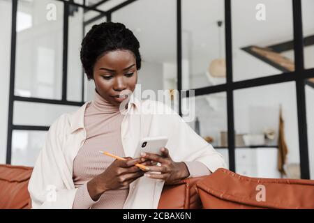 Immagine di una giovane donna africana piuttosto concentrata seduta sul divano in ambienti interni a casa mentre si utilizza il telefono cellulare Foto Stock