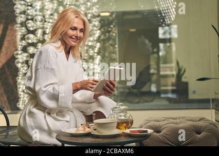 Signora affascinante in accappatoio bianco seduta vicino alla scrivania di vetro Foto Stock